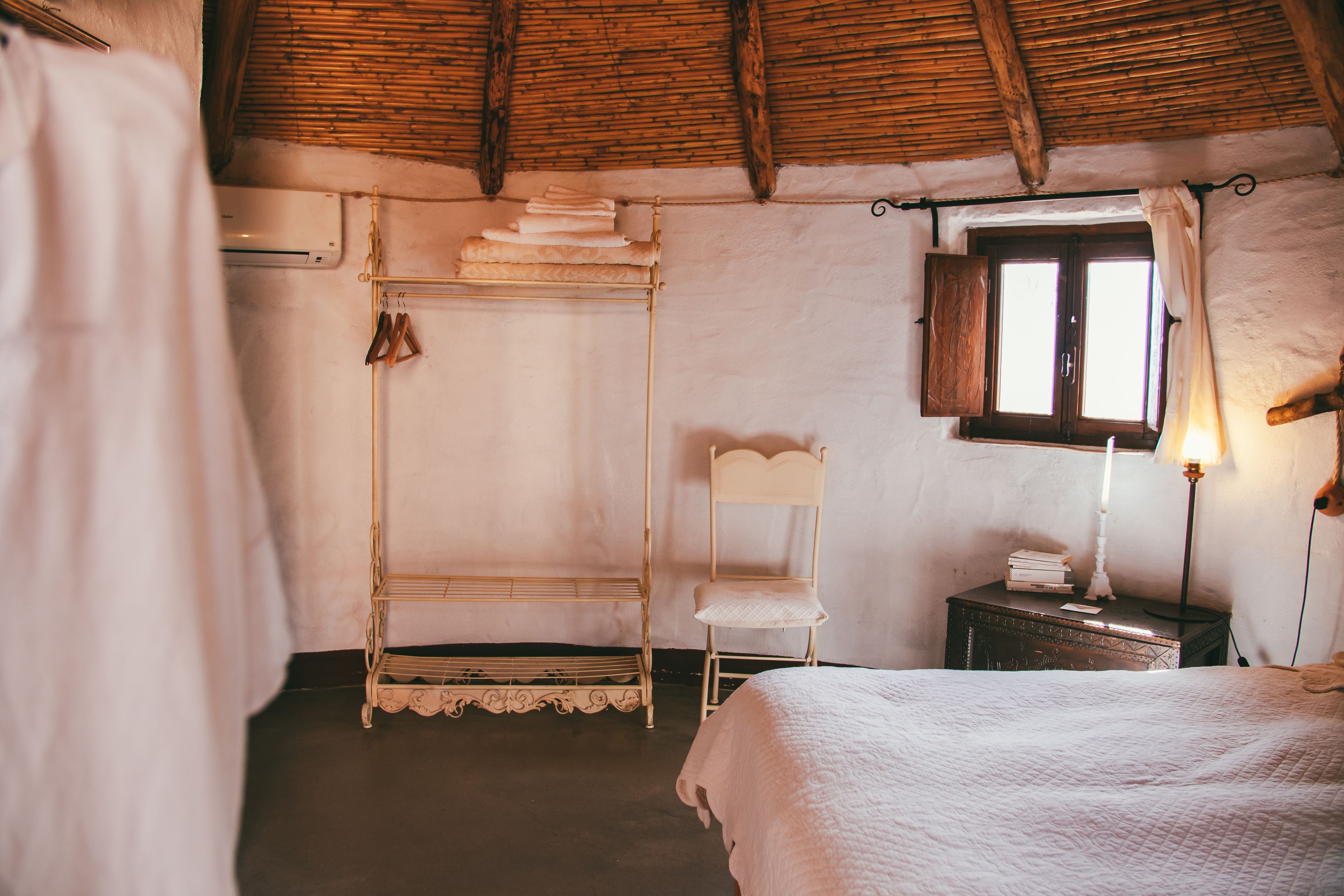 A rustic bedroom with a four-poster bed, a small bedside table with a lamp, a chair, and a window with curtains.