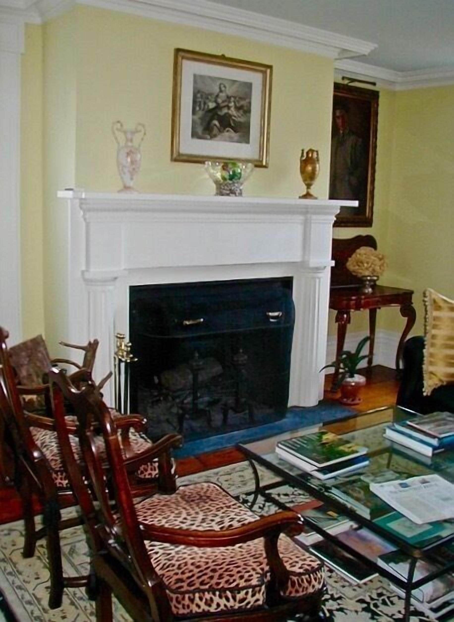 A room with a fireplace, wooden chairs, a glass table with books, and a framed picture on the wall.