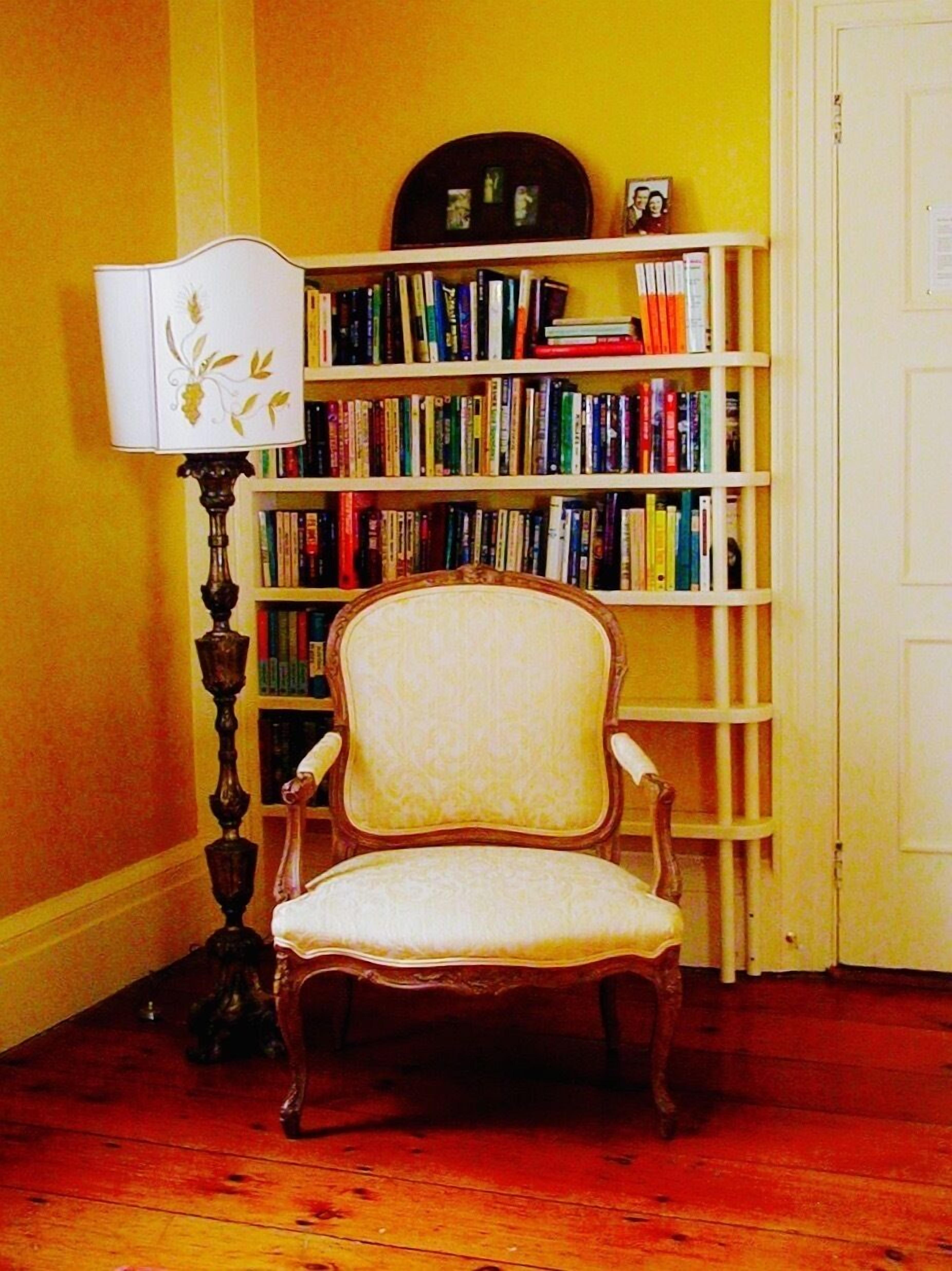 Ein Zimmer mit Holzboden, einem weißen Stuhl mit gepolstertem Sitz, einer hohen Stehlampe mit dekorativer Schirm und einem Bücherregal voller Bücher.