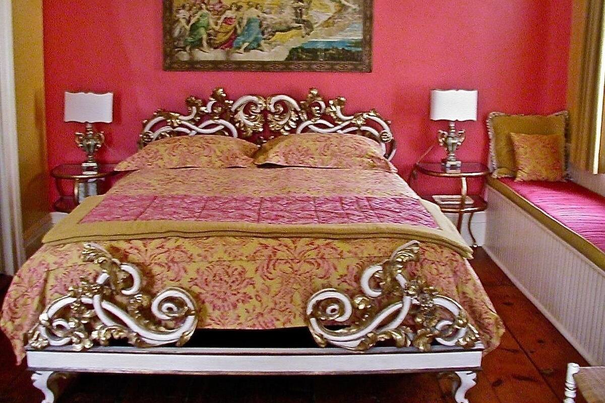 Un lit doté d’une tête de lit et d’un pied de lit en métal décoratif, flanqué de deux tables de chevet avec des lampes, dans une pièce dont un mur est rouge et où un tableau est accroché au-dessus du lit.