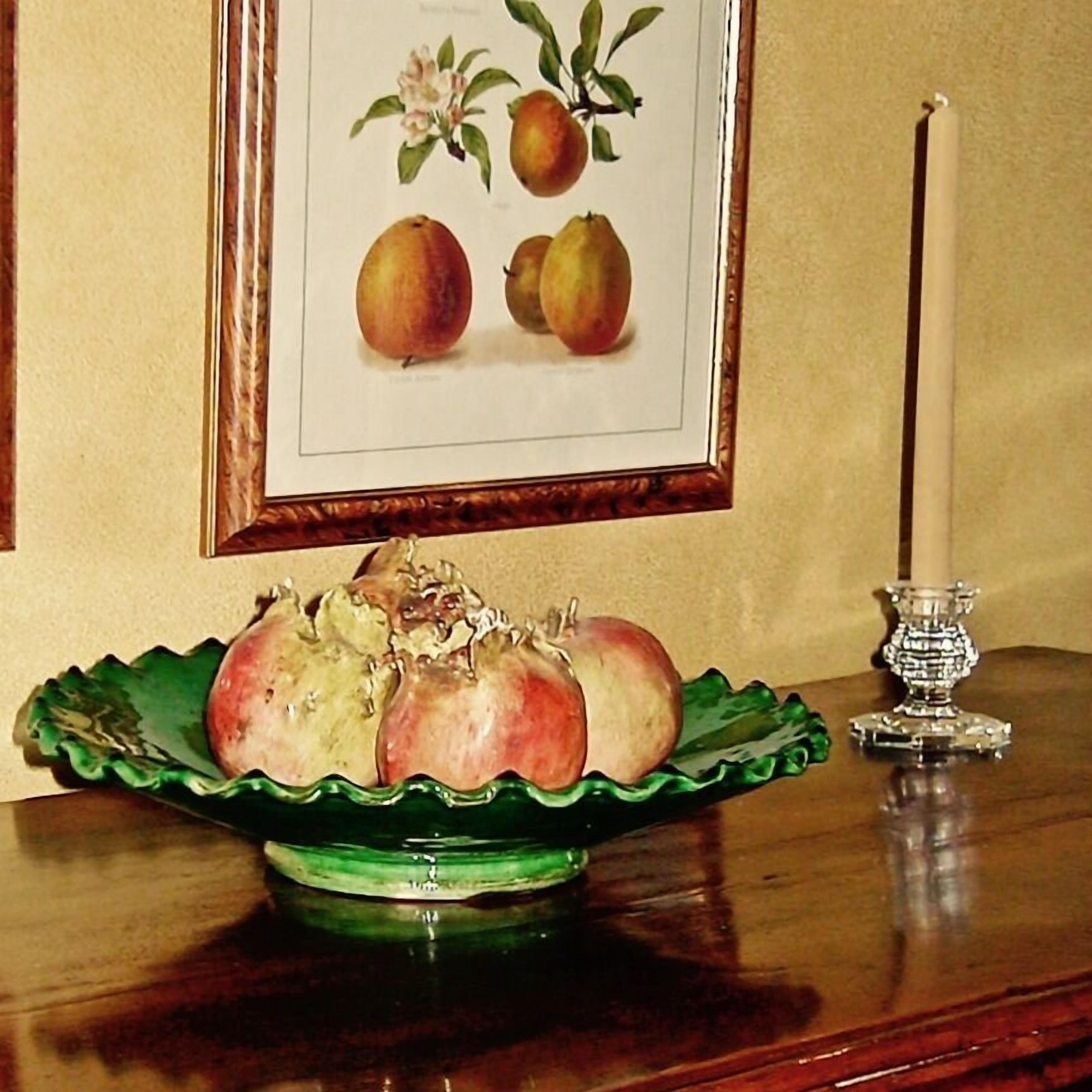 A green bowl with pomegranates on a wooden table, a framed illustration of fruits above it, and a candle holder to the right.