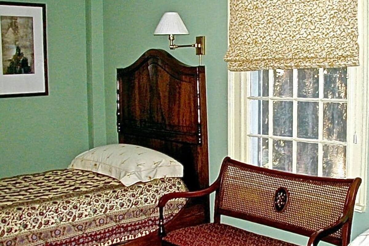 Une chambre à coucher avec un lit en bois, une chaise, une fenêtre agrémentée d’une lambrequin à motifs et un tableau encadré accroché au mur.