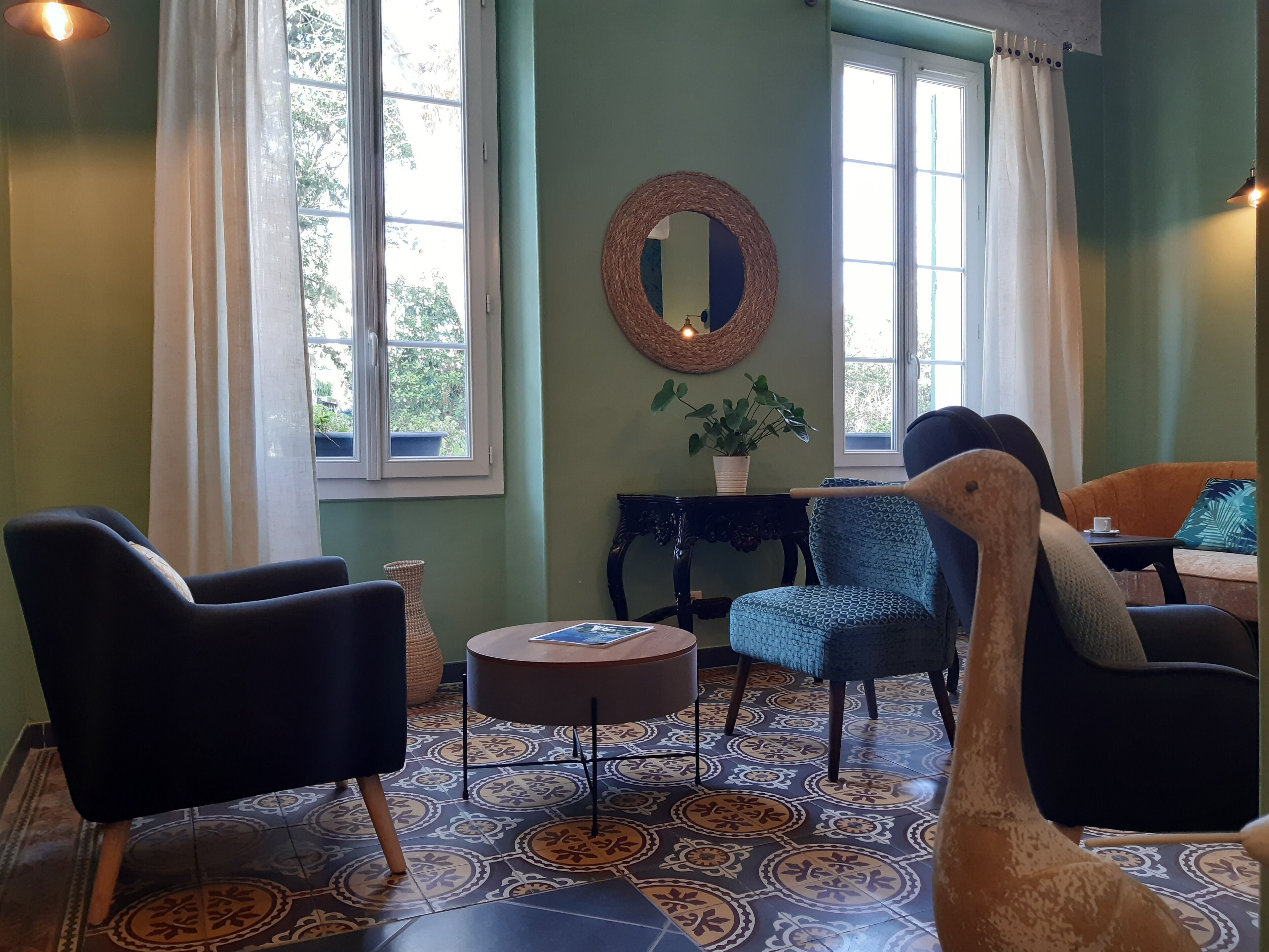 A room with two black armchairs, a round coffee table, a wicker mirror, a potted plant, and a patterned floor.
