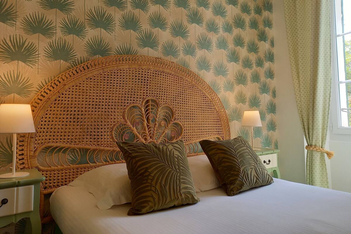 A room with a large bed, two bedside lamps, a rattan headboard, and patterned wallpaper.