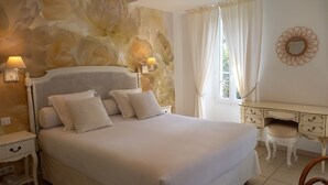A bedroom with a large bed, a desk, and a chair. There is a wall with a floral design, a window with curtains, and two wall-mounted lamps. - Hôtel Notre Dame (Collobrieres)