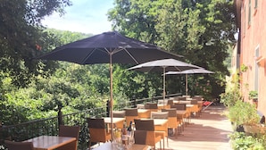 An outdoor patio with wicker tables and chairs, shaded by large umbrellas, surrounded by lush greenery. - Hôtel Notre Dame (Collobrieres)