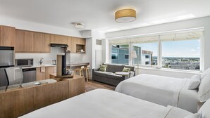 Habitación de hotel moderna con kitchenette, sofá, cama y un amplio ventanal con vista a la ciudad.