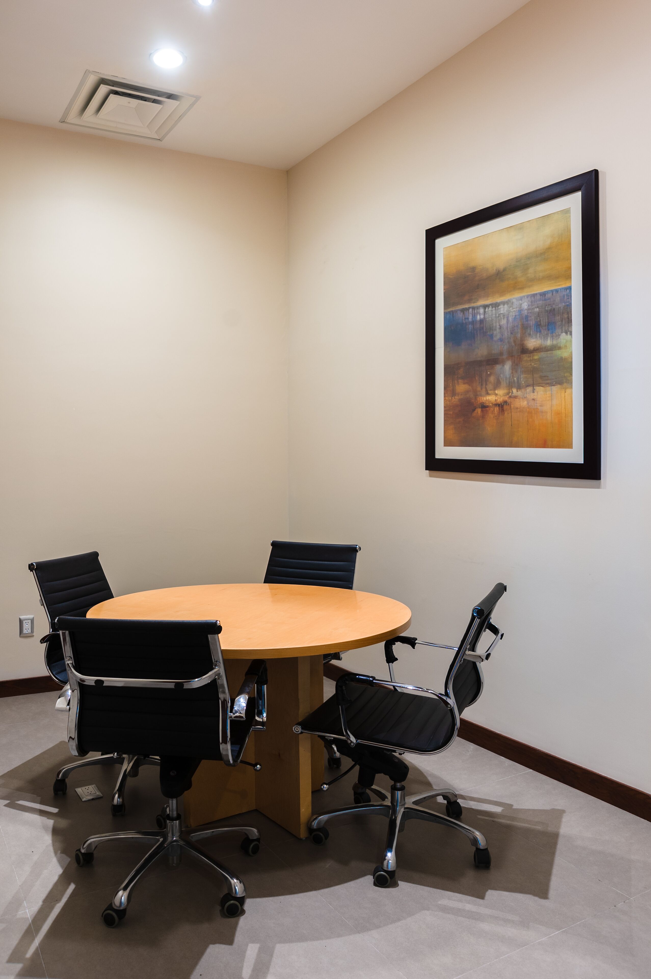 Uma sala de conferências com uma mesa redonda, quatro cadeiras de escritório pretas e um quadro abstrato emoldurado na parede.