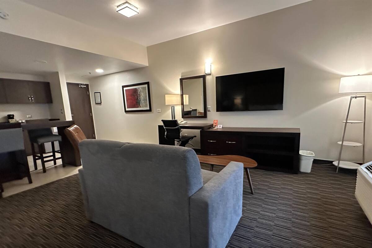 Ein Hotelzimmer mit grauem Sofa, einem Schreibtisch mit Stuhl, einem an der Wand befestigten Fernseher und einer kleinen Küchenzeile.