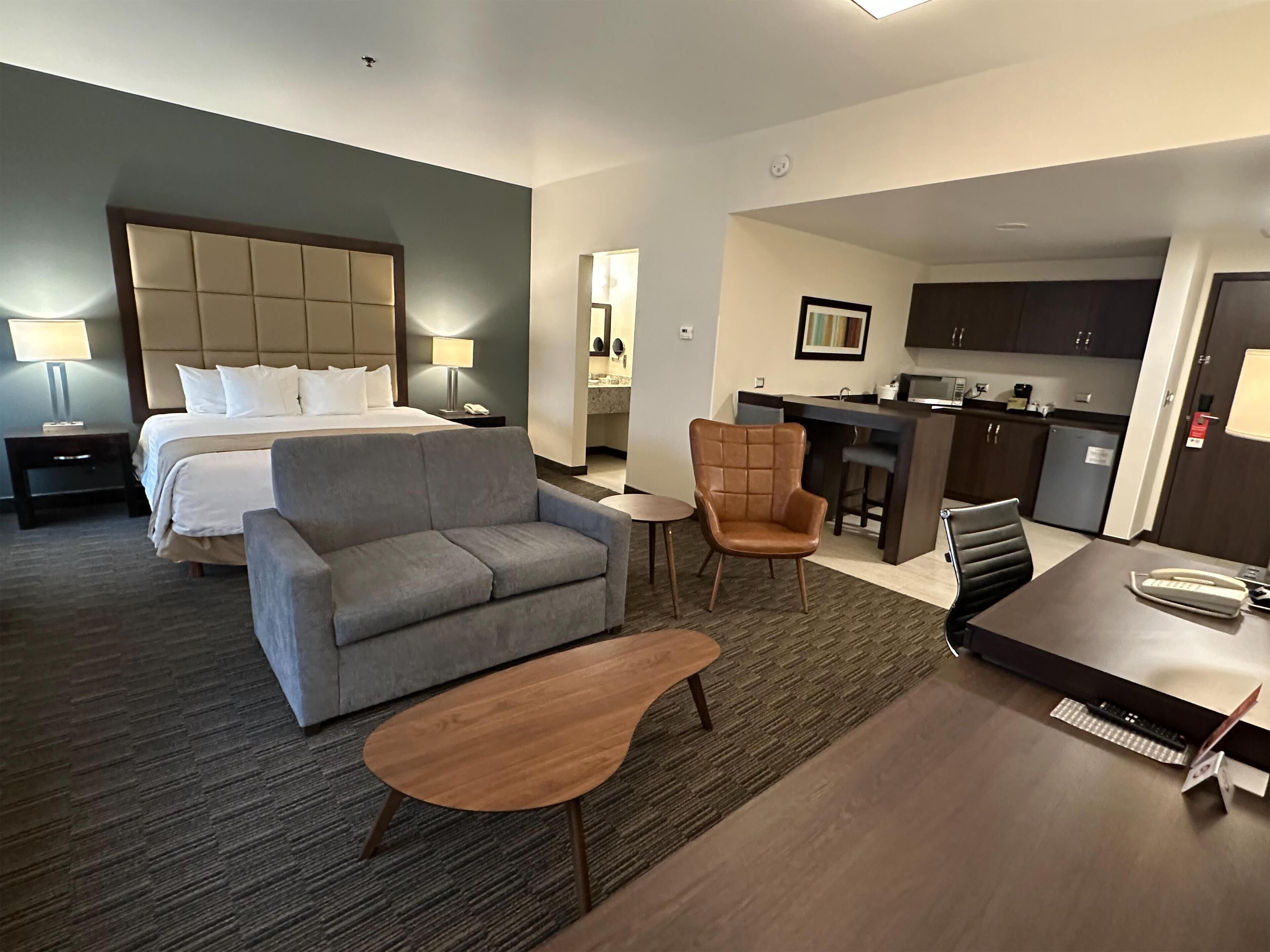 A hotel room with a bed, a grey sofa, a wooden coffee table, a brown armchair, and a small kitchen area with a bar and cabinets.