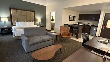 Una habitación de hotel con una cama, un sofá gris, una mesa de centro de madera, un sillón marrón y una pequeña cocina con barra y armarios.