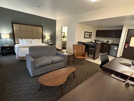 Una habitación de hotel con una cama, un sofá gris, una mesa de centro de madera, un sillón marrón y una pequeña cocina con barra y armarios.
