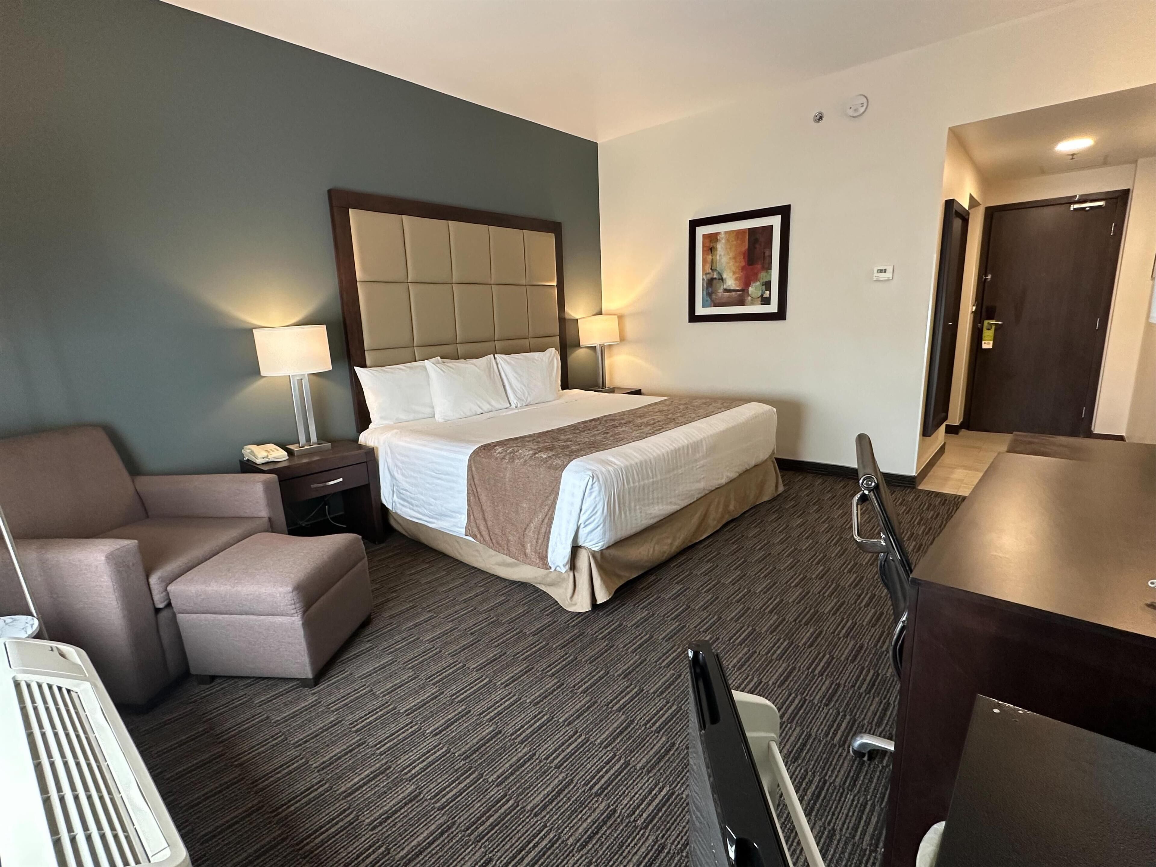 A hotel room with a bed, a desk, a chair, a lamp, and a heater.