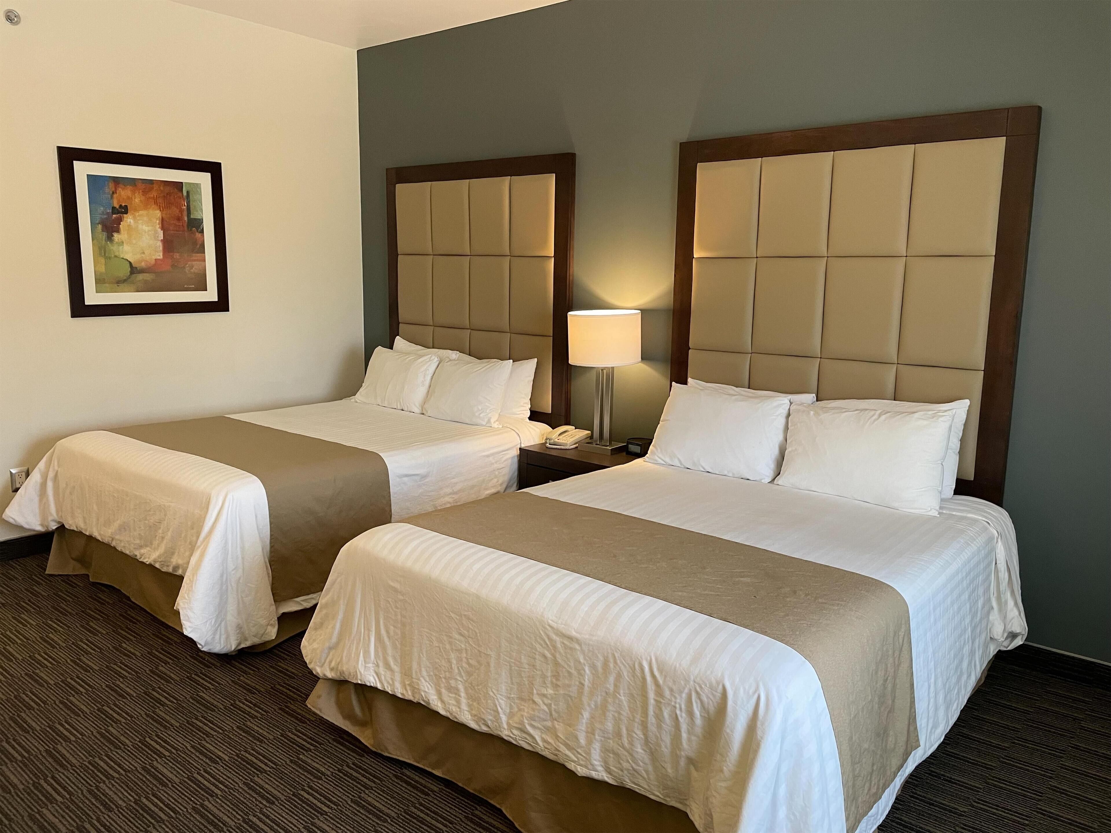 Habitación de hotel con dos camas, cada una con cabecero, mesita de noche con lámpara y un cuadro en la pared.
