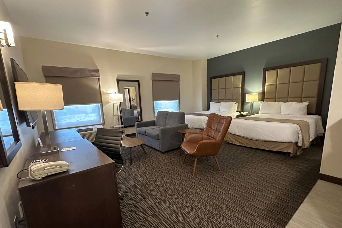 Ein Hotelzimmer mit Schreibtisch, zwei Sesseln, einem braunen Ledersessel, einem Bett mit zwei Kissen und einem großen Fenster.