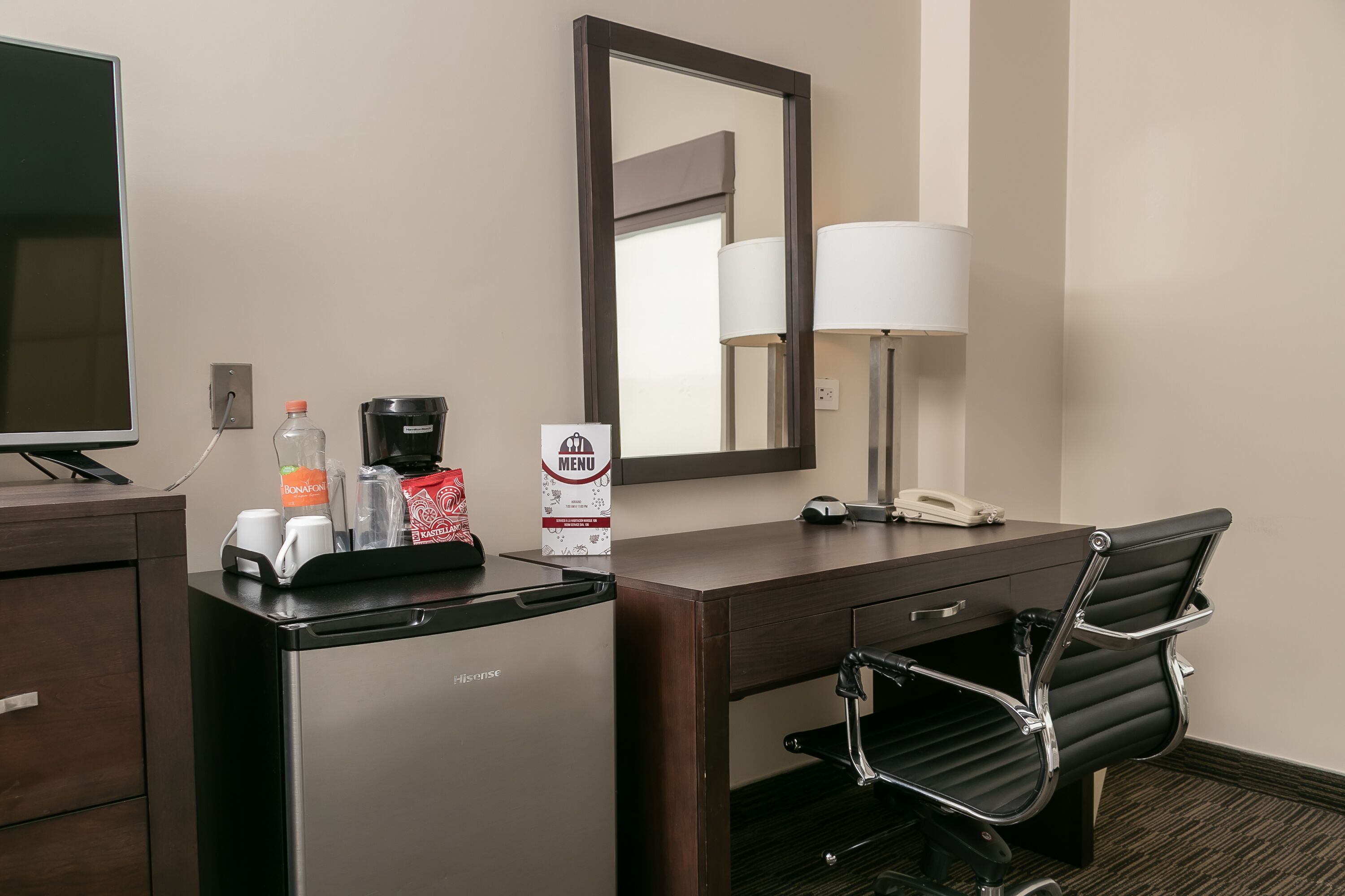Une chambre d’hôtel équipée d’un bureau, d’une chaise, d’une machine à café, d’une bouteille, d’un verre, d’un menu, d’une lampe et d’un miroir.
