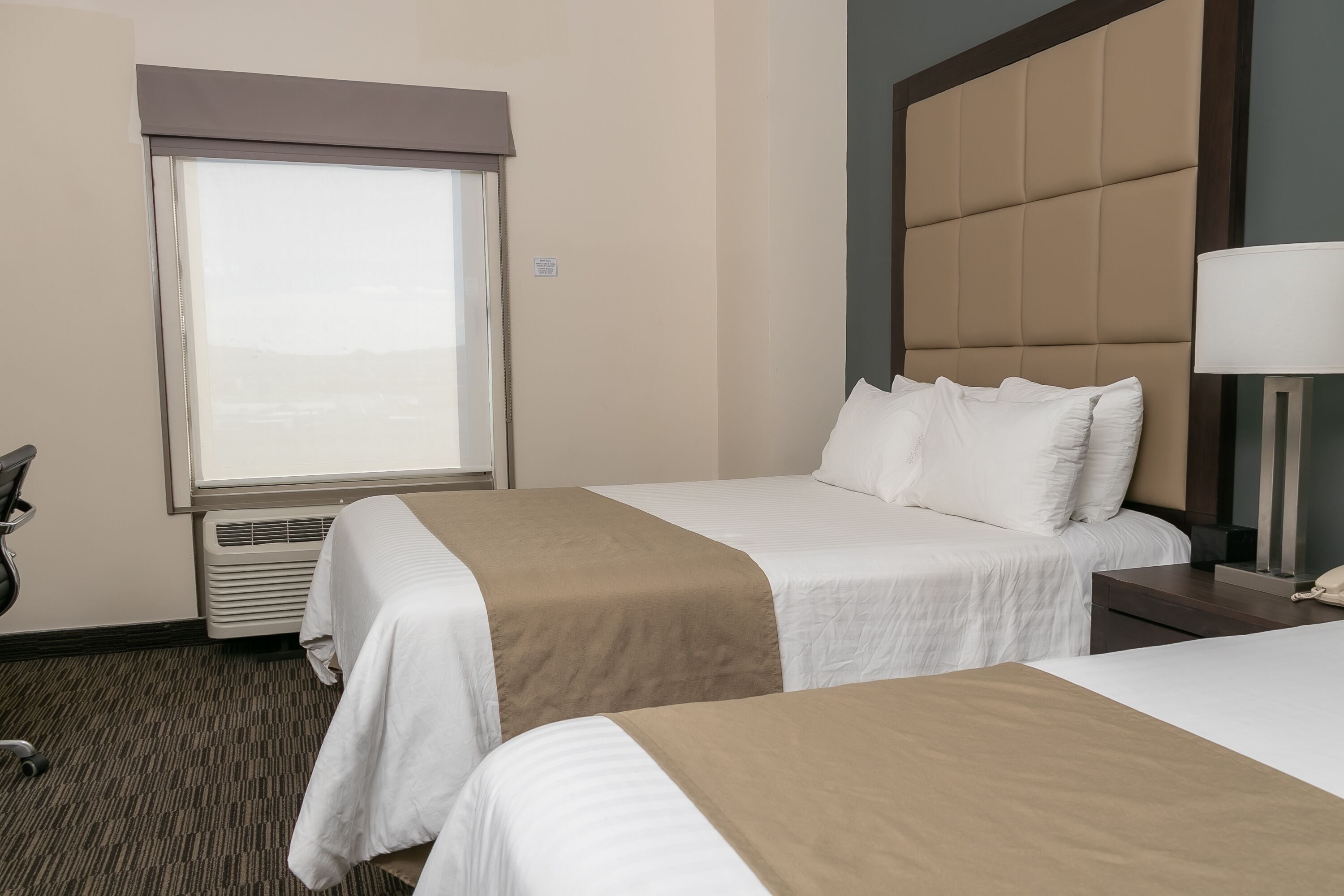 A hotel room with two beds, a large window, a headboard, and a bedside lamp.
