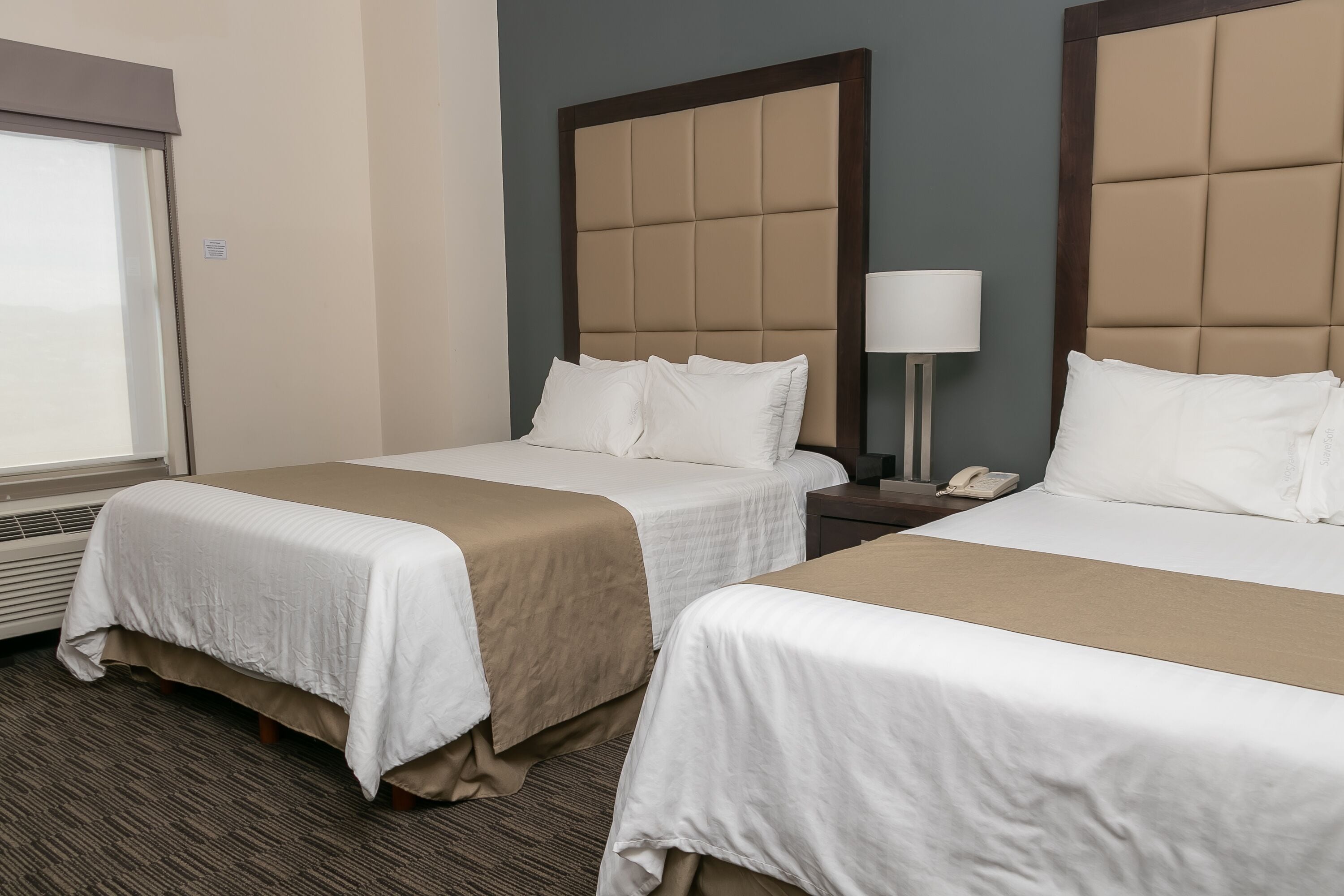 Een hotelkamer met twee bedden, een nachtkastje met een lamp en een raam met jaloezieën.
