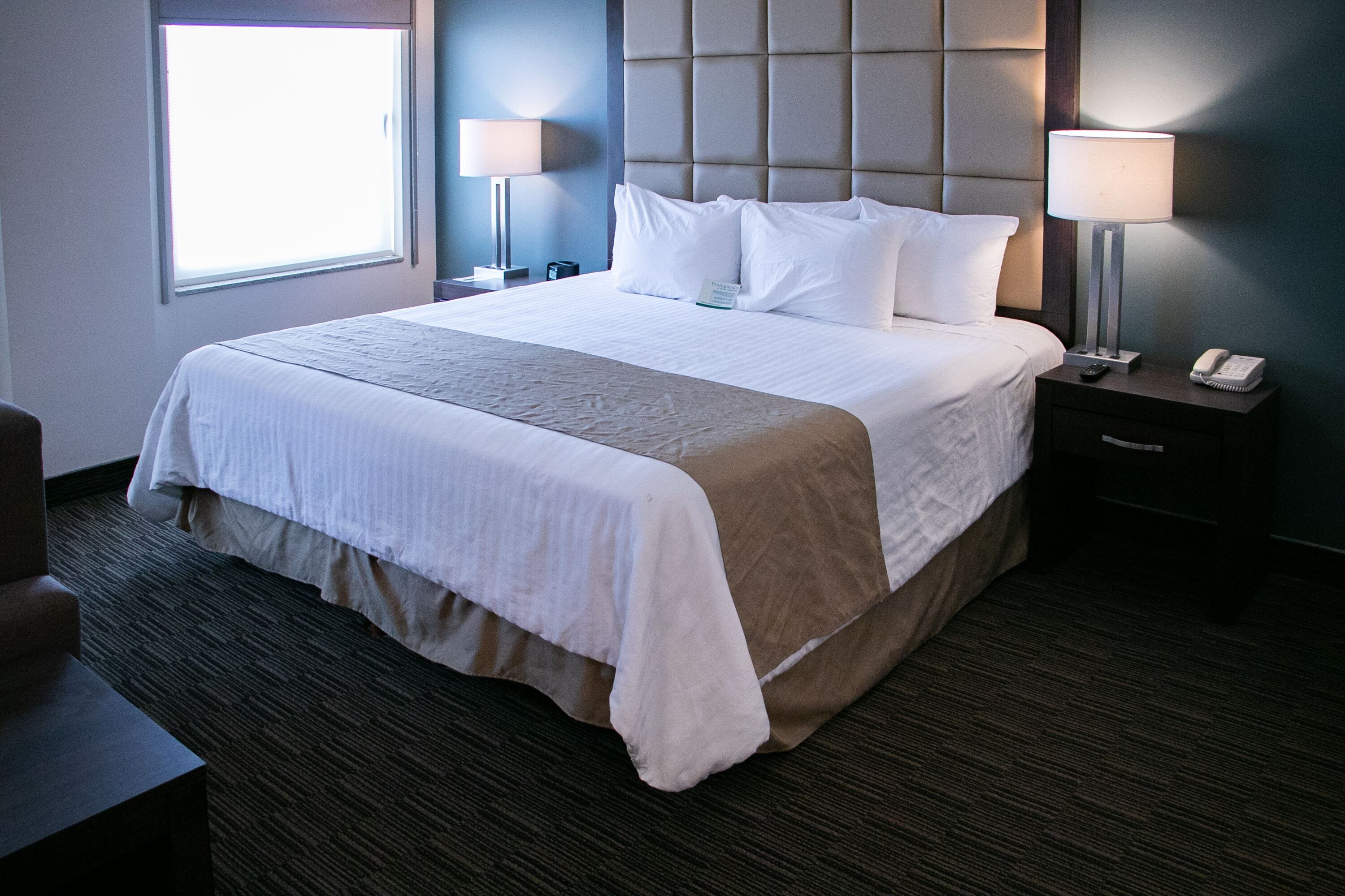 A hotel room with a large bed, two bedside lamps, a nightstand with a phone, and a window providing natural light.