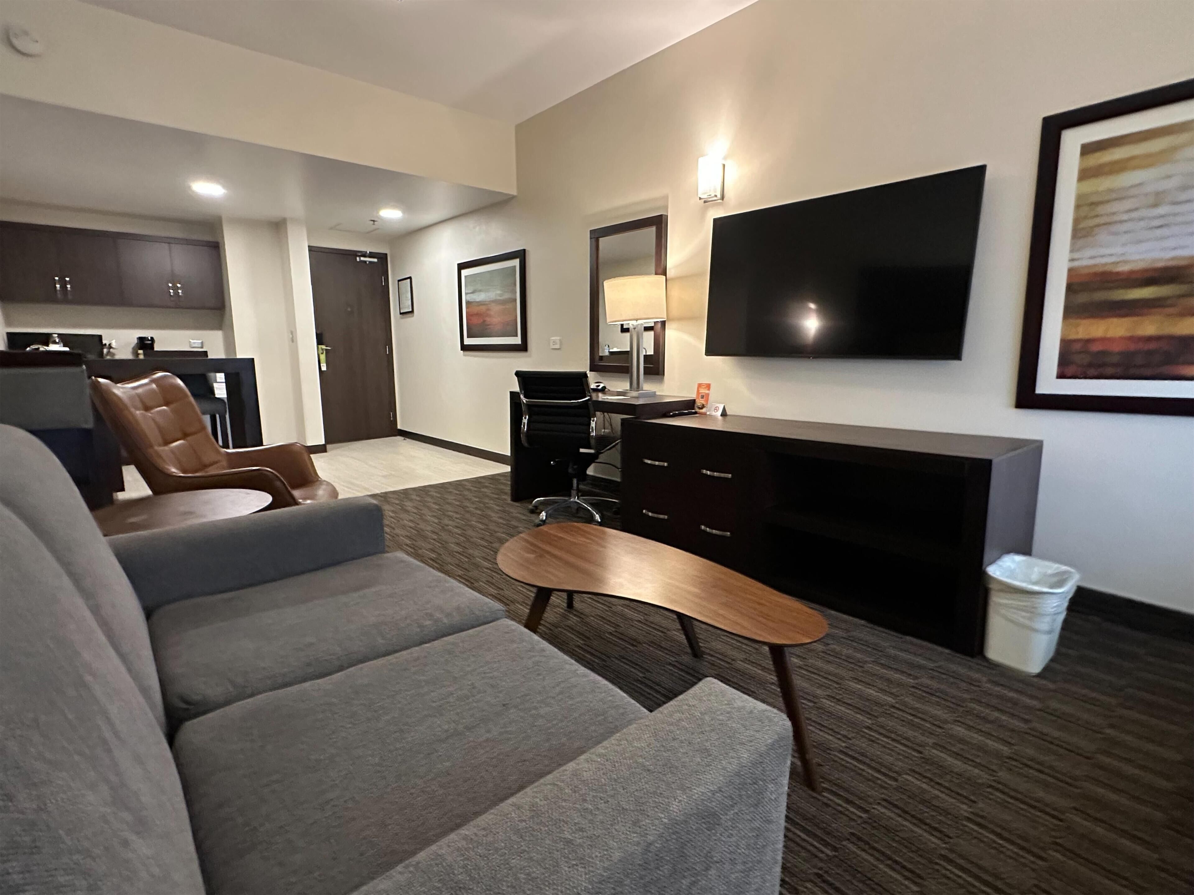 Una habitación de hotel moderna con un sofá gris, una mesa de centro de madera, un televisor de pantalla plana montado en la pared, un escritorio pequeño con una silla y una zona de cocina al fondo.