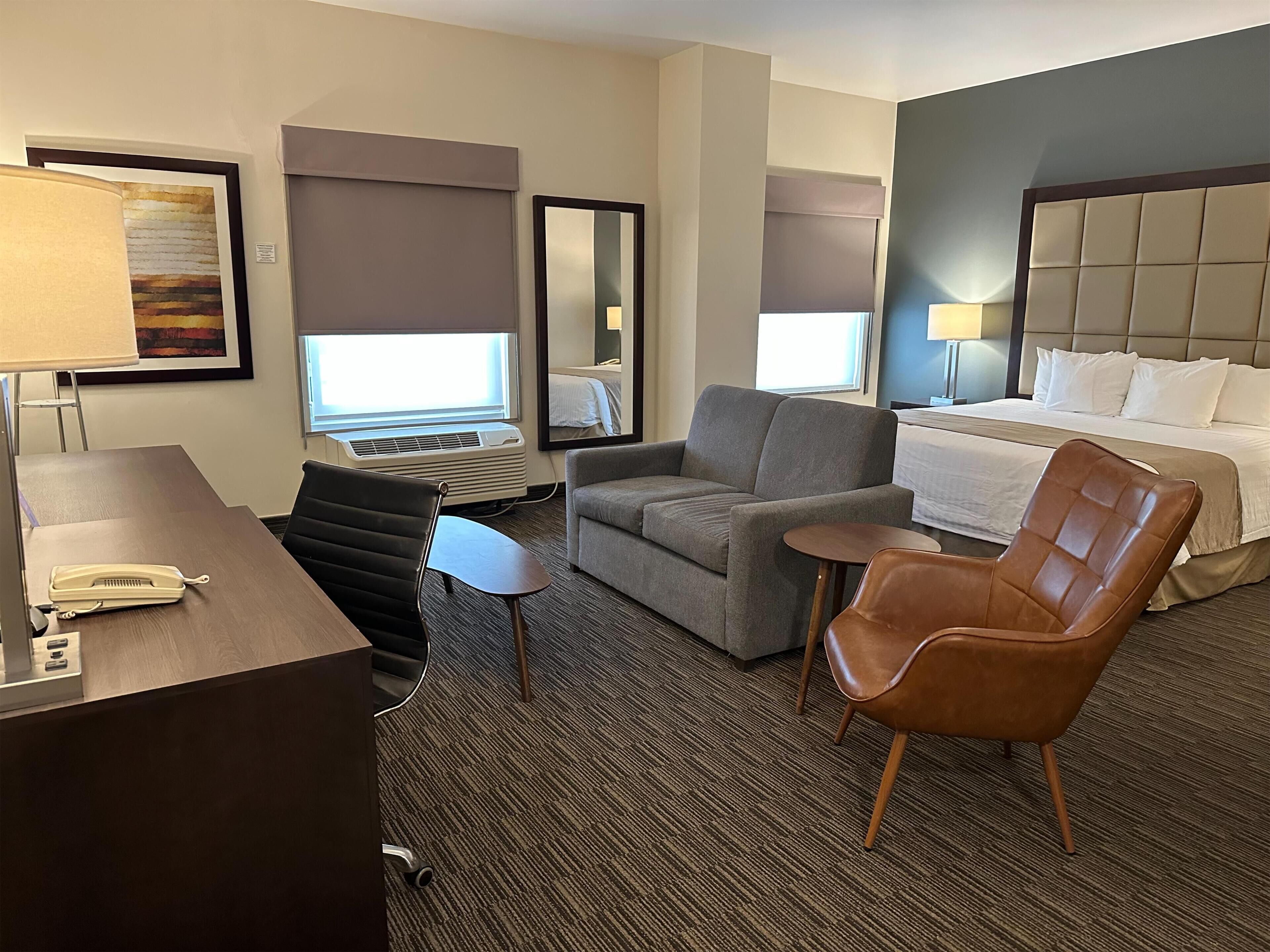 Una habitación de hotel con una cama, un escritorio con teléfono, un sillón gris y un sillón de cuero marrón, junto a una pequeña mesa.
