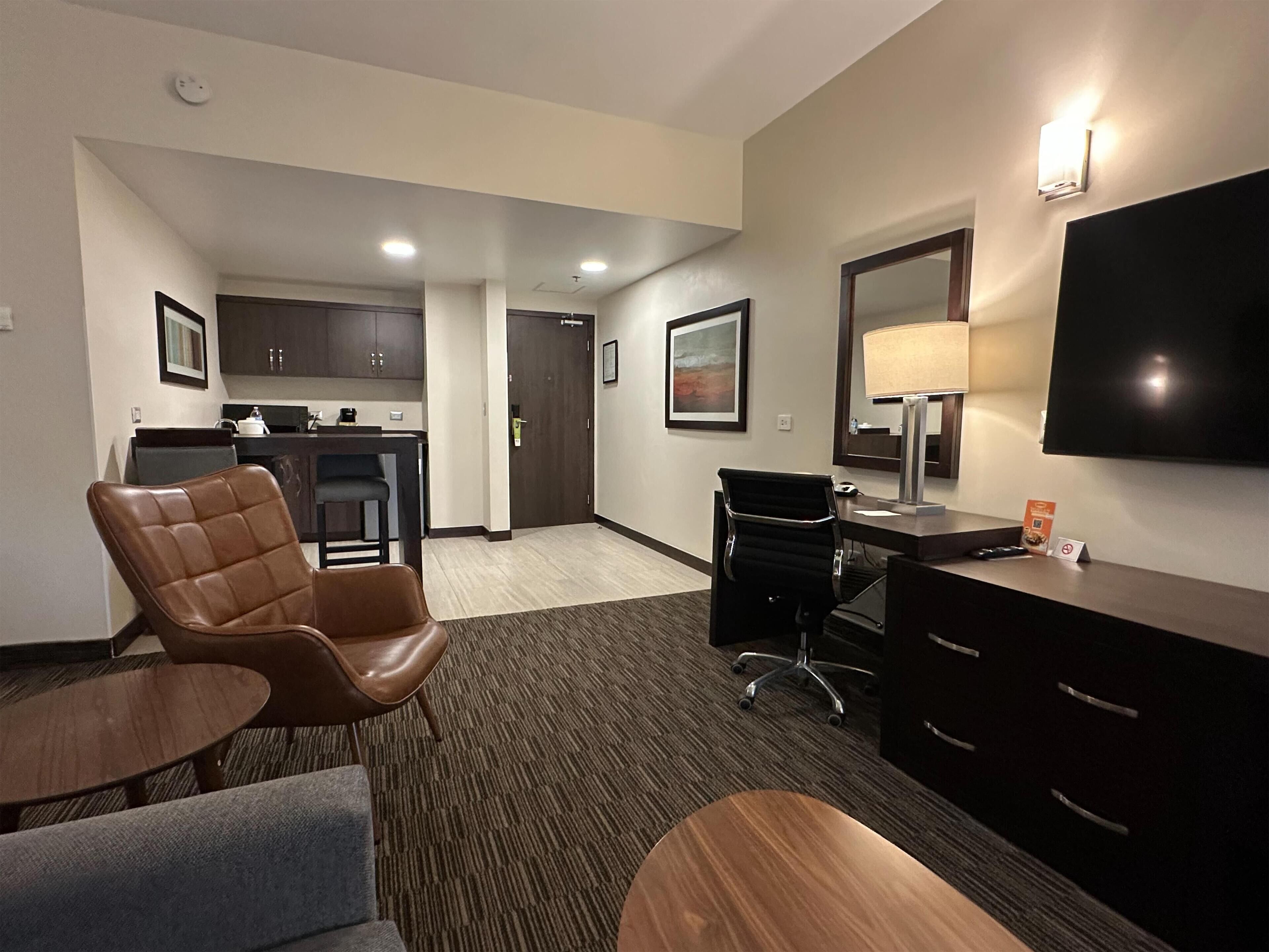 A hotel room with a brown leather chair, a desk with a lamp, a flat-screen TV, a small table, and a kitchenette in the background.