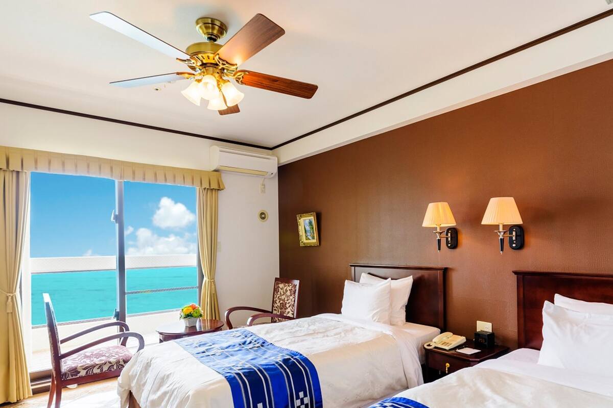 Een hotelkamer met twee bedden, een plafondventilator, uitzicht op de oceaan en een balkon met een stoel.