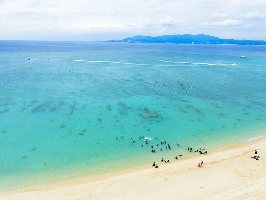 Una playa con aguas turquesas cristalinas, una orilla arenosa y montañas a lo lejos.