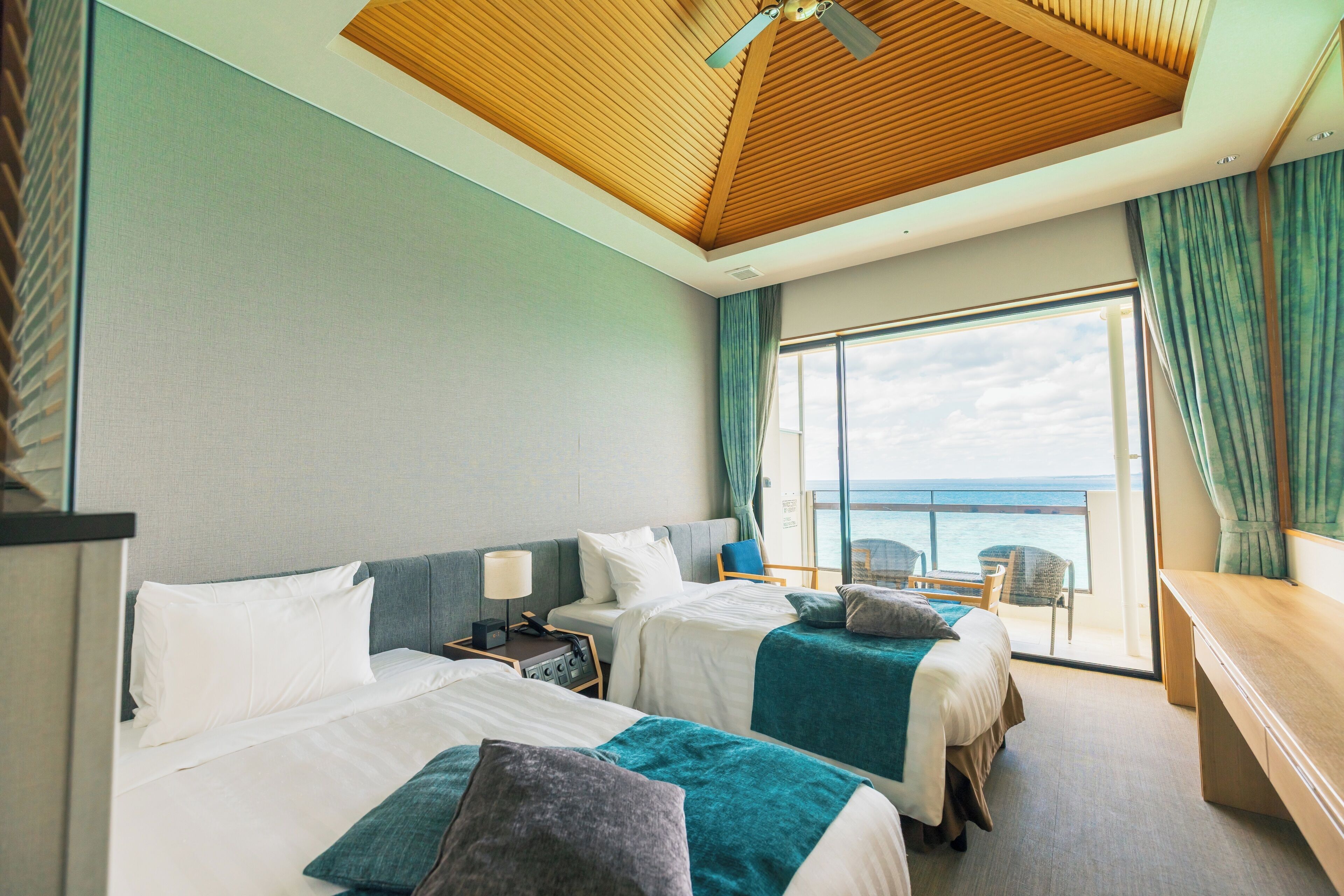 Ein Hotelzimmer mit zwei Betten, einer Holzbalkendecke, einem Balkon mit Meerblick und einem Schreibtisch.