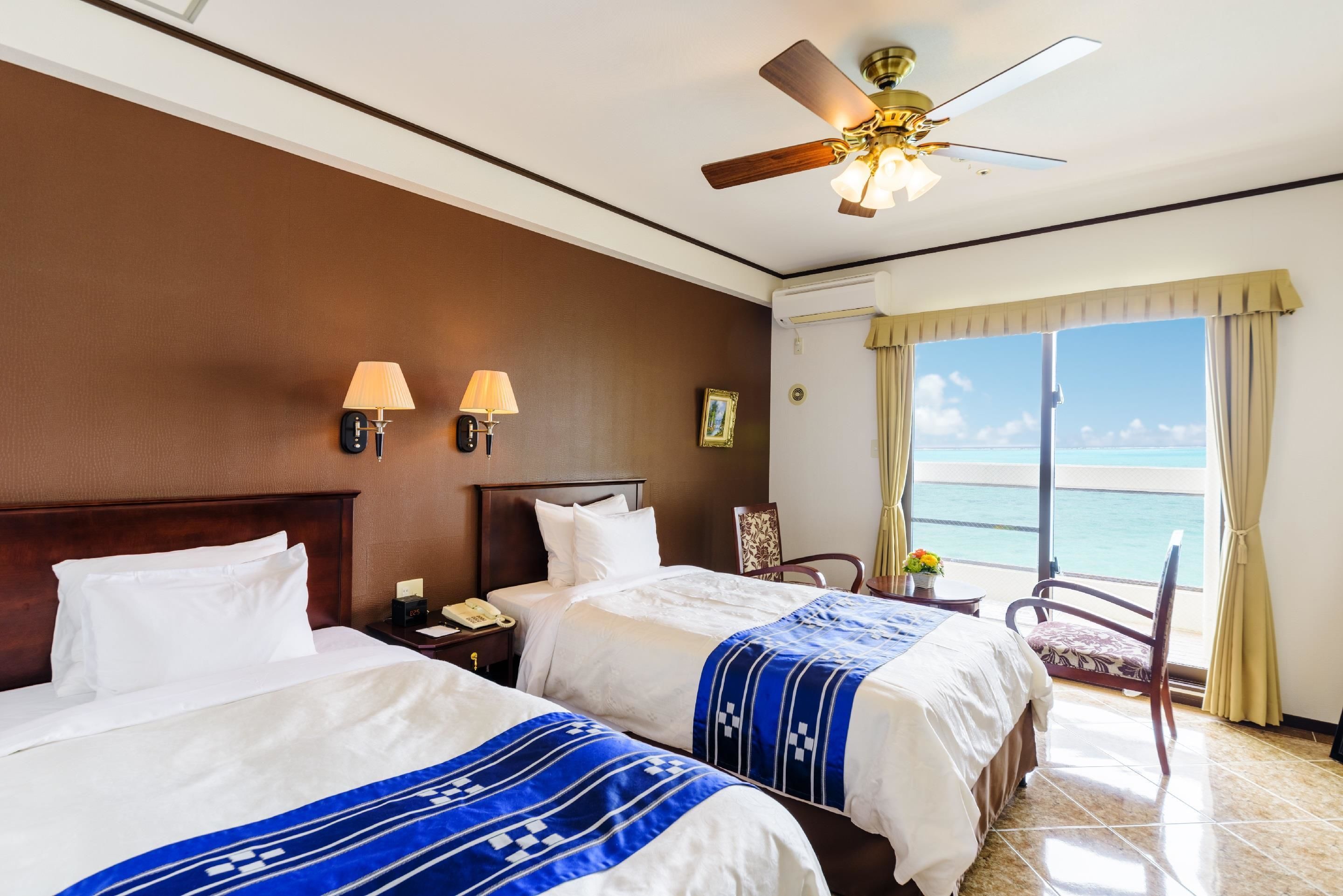 A hotel room with two beds, a ceiling fan, a nightstand with a lamp, a chair, and a view of the ocean.