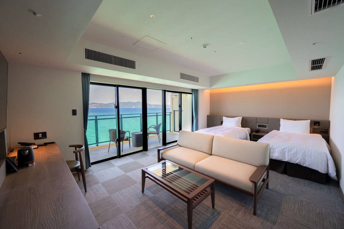 Hotelkamer met een groot bed, een bank, een glazen salontafel en uitzicht op zee door grote ramen.