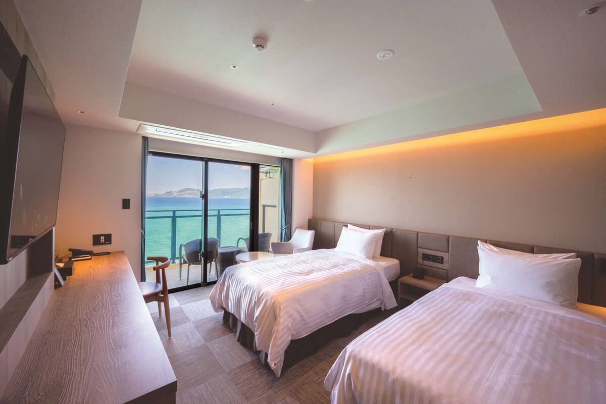 Een hotelkamer met twee bedden, een bureau, een stoel en een balkon met uitzicht op zee.