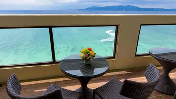 Un balcon avec une table en verre et des chaises en osier, offrant une vue sur une mer turquoise et des montagnes lointaines.