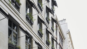 Un immeuble à plusieurs étages doté de grandes fenêtres, dont certaines sont surmontées d’auvents noirs et ornées de plantes en pot. Un homme en costume se tient sur le trottoir.