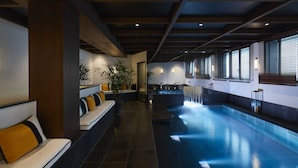 Un espace piscine intérieur moderne, avec des sièges le long des murs, un élément décoratif aquatique et un plafond avec des poutres apparentes.