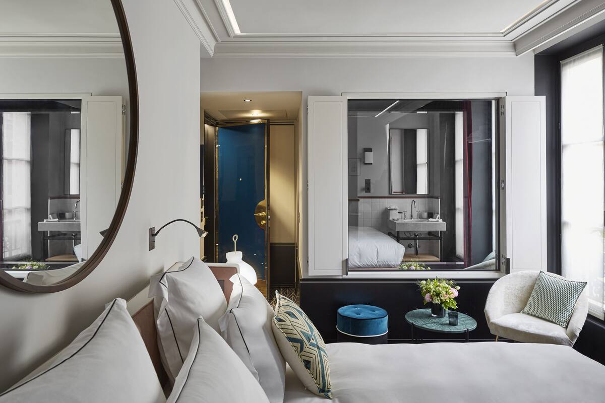 A modern hotel room with a large bed, a round mirror, a small round table with a vase, and a view of the bathroom through a large window.