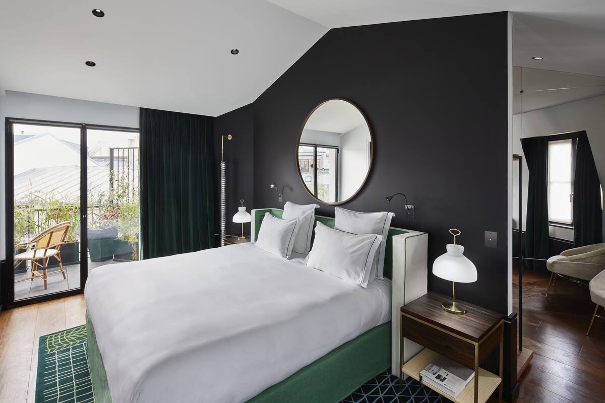 A modern hotel room with a large bed, a round mirror, a nightstand with a lamp, and a view of the outdoors through a sliding door.
