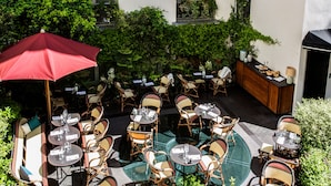 Une terrasse extérieure avec des tables rondes et des chaises en osier, entourée de verdure et bordée d’un bâtiment blanc.