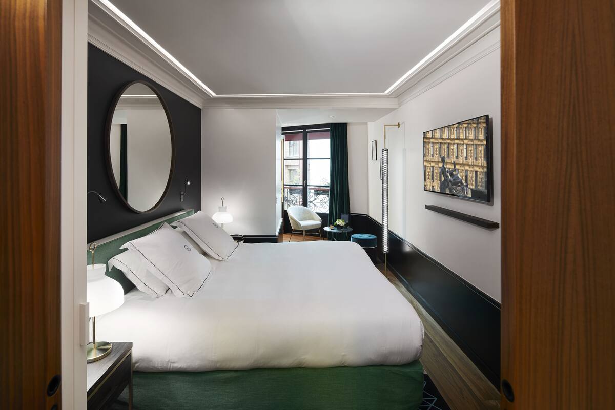 A hotel room with a large bed, a round mirror, a chair, a desk with a lamp, and a painting on the wall.
