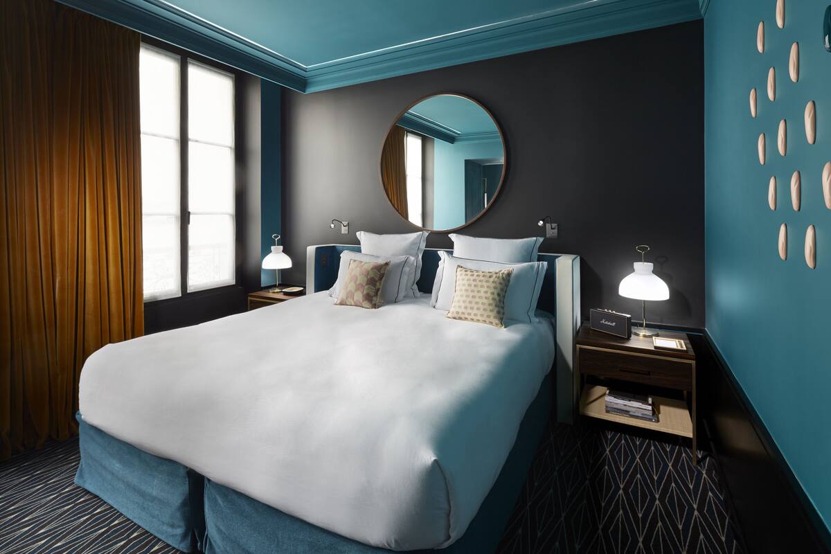 A hotel room with a large bed, two bedside tables, a round mirror, and a view of the city through the window.
