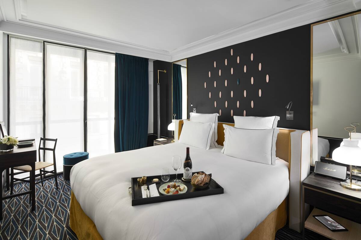 A hotel room with a large bed, a bedside table with a lamp, a desk with a chair, and a window with curtains.
