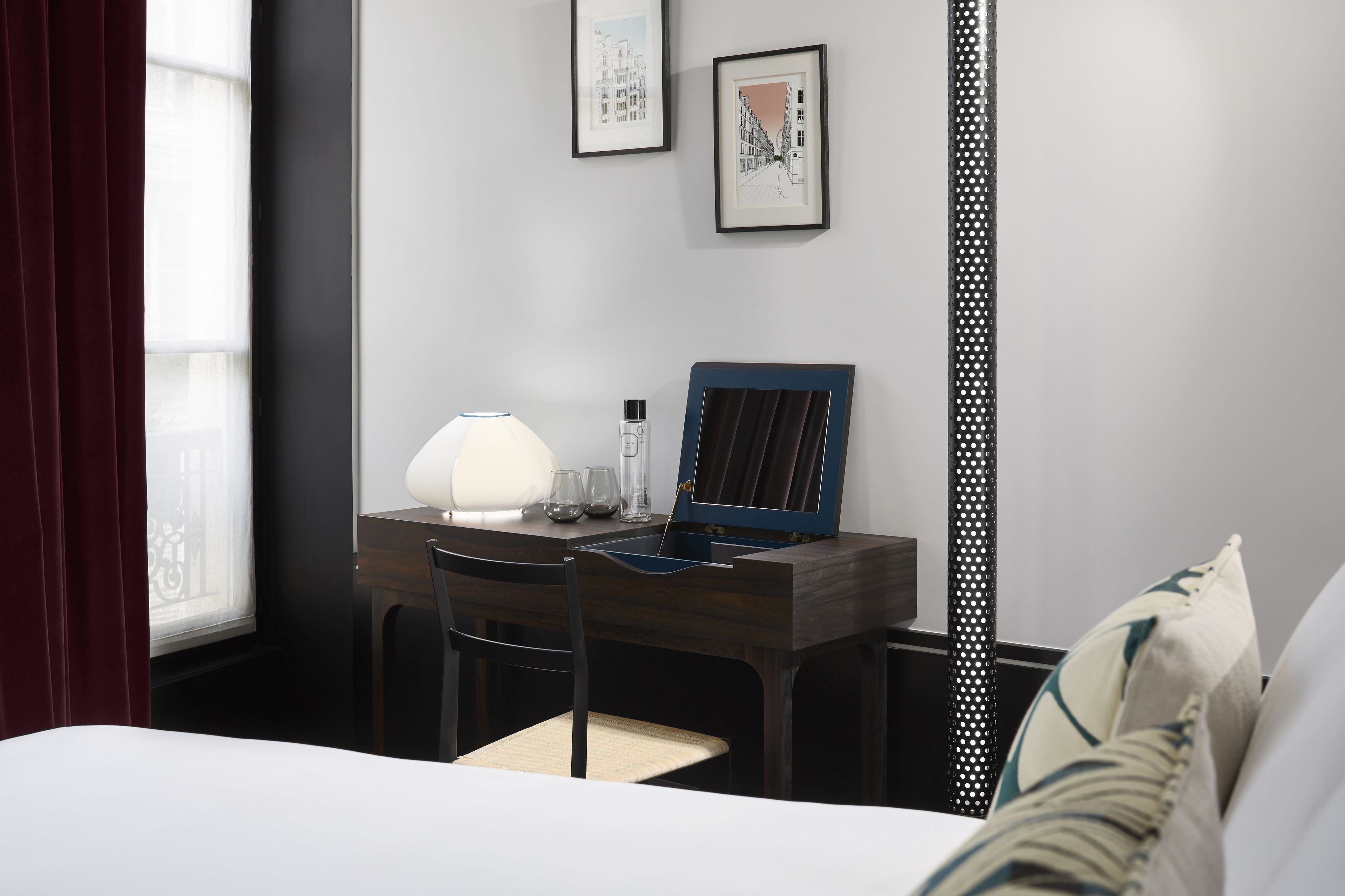 Habitación de hotel con escritorio, silla y cama. En la pared, hay cuadros enmarcados, una lámpara sobre el escritorio y una ventana con cortinas.