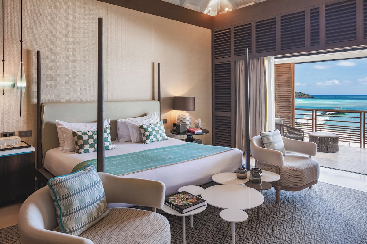 A modern hotel room with a large bed, a balcony with a view of the sea, and a seating area with a small table.