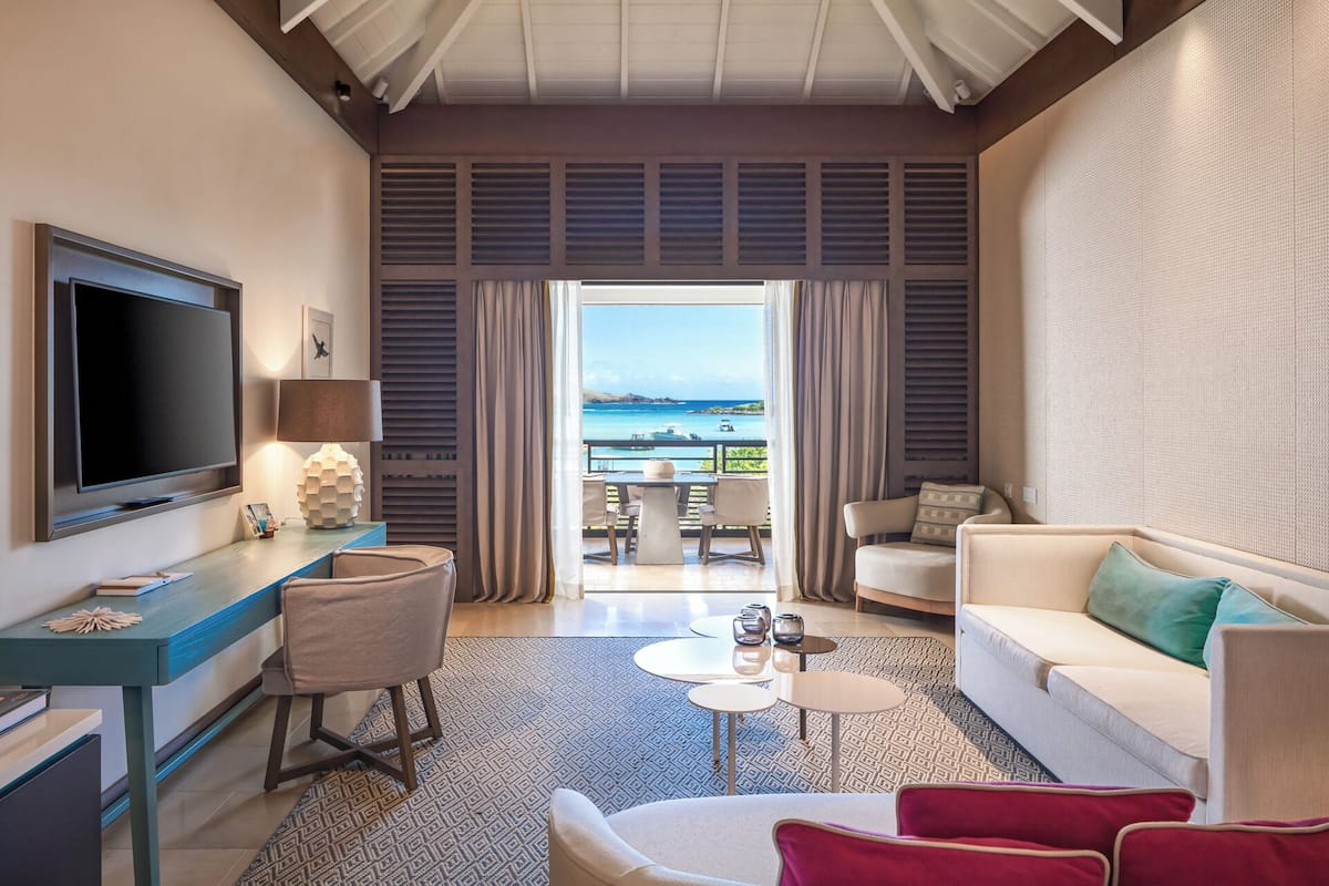 A modern hotel room with a flat-screen TV, a desk, a sofa, and a balcony with a view of the sea.