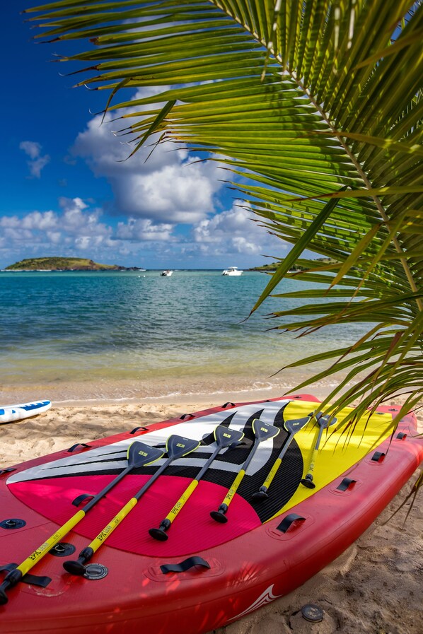Um caiaque vermelho com remos e uma prancha de stand-up paddle amarela numa praia arenosa com água cristalina e uma ilha ao longe.