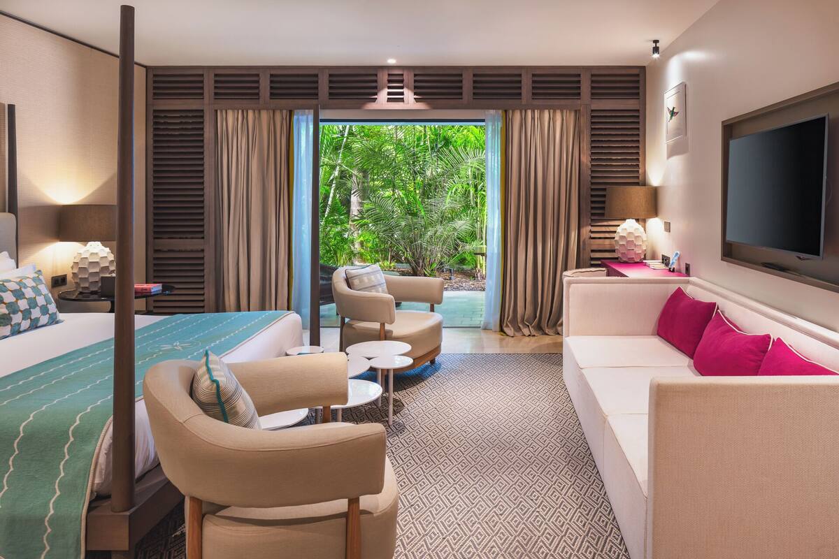 A hotel room with a bed, a sofa, a chair, a small table, a TV, and a view of greenery outside.