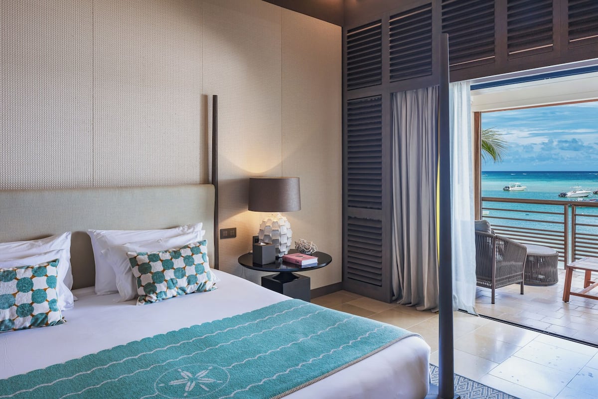 A hotel room with a bed, a nightstand, a lamp, a view of the ocean, and a balcony with chairs.