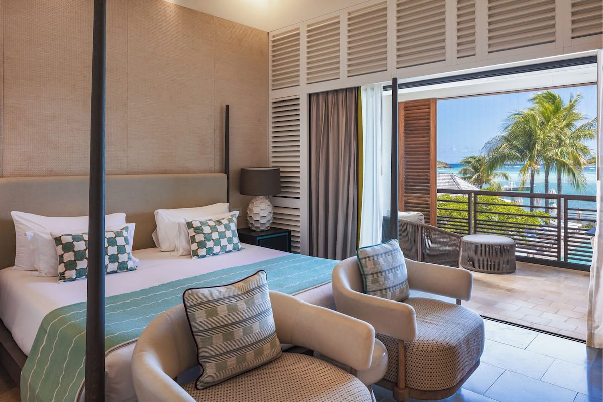 A hotel room with a bed, two armchairs, a small table, a nightstand with a lamp, and a balcony with a view of the ocean and palm trees.