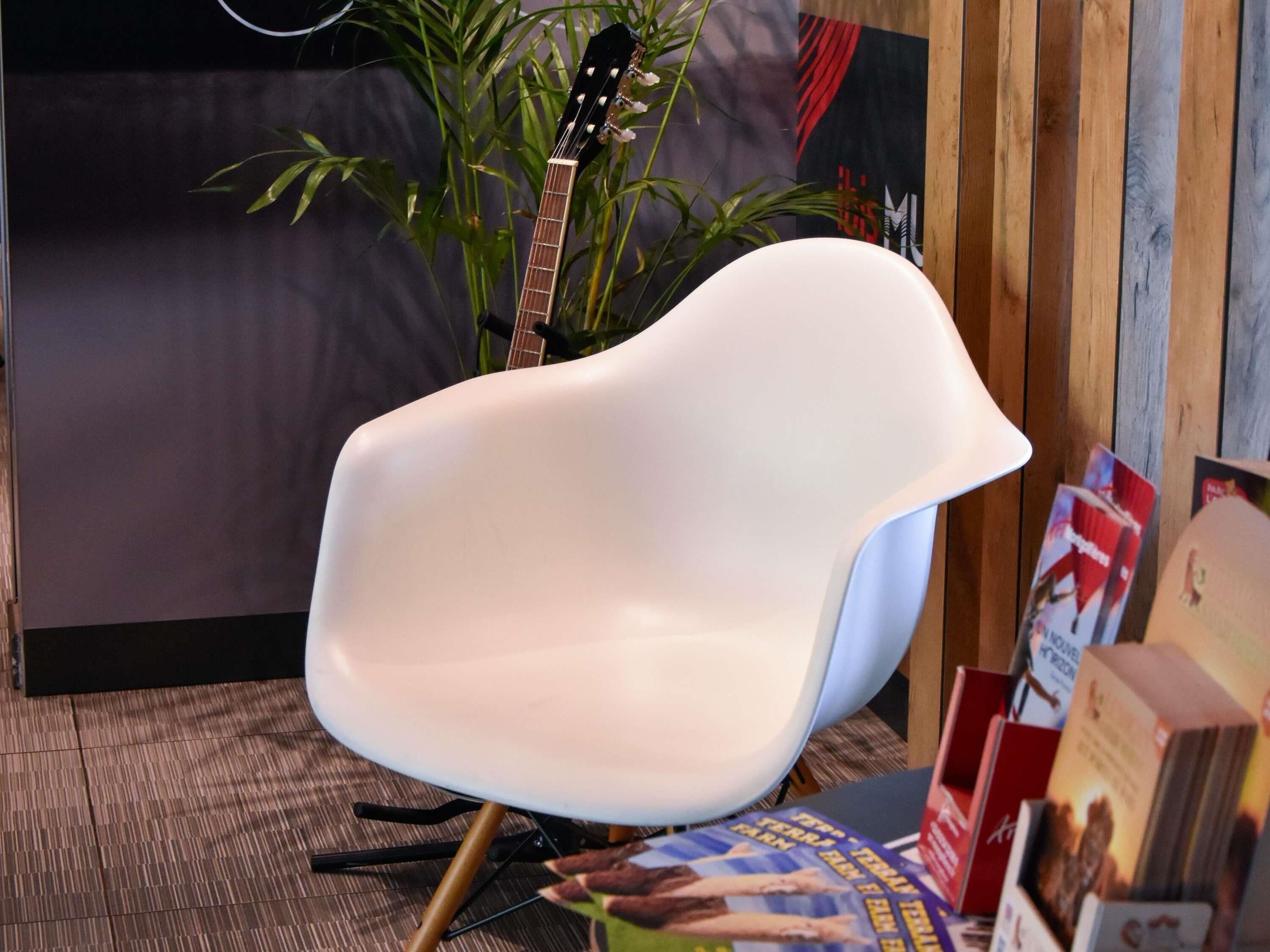 Una silla blanca con respaldo curvo, una guitarra apoyada contra una pared y una planta al fondo.
