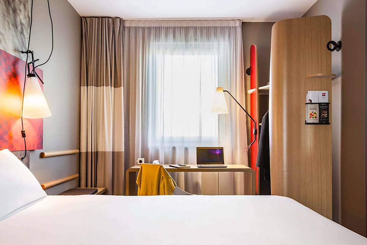 A hotel room with a bed, a desk with a laptop, a chair, a lamp, and a window with curtains.